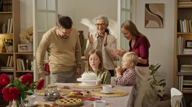 Caucasian girl blowing birthday cake candles while multigenerational family claps and father gives gift box. Clip works well for family celebration commercials and holiday promotional campaigns