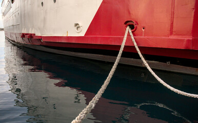 Mooring rope extending through vessel hawsehole on red and white ship hull in calm morning harbor. © YuliiaKettu