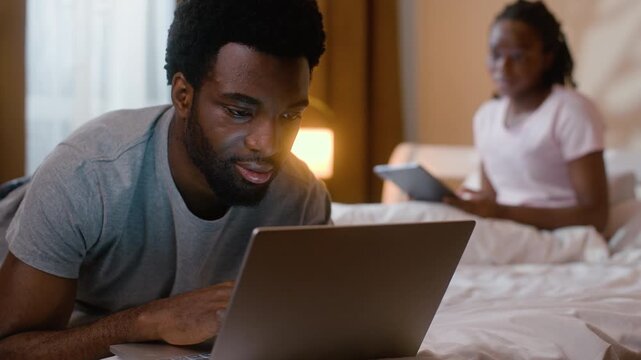 Happy African American family relaxing in bed using laptop digital tablet at night husband wife scrolling social media online device ethnic couple marriage gadget addiction weekend home bedroom indoor