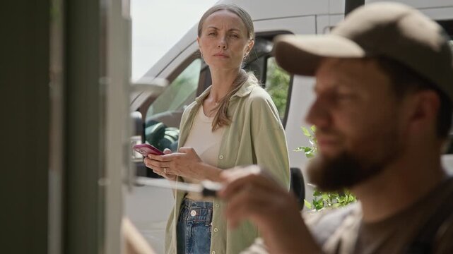 Caucasian handyman fixing door lock with screwdriver while middle aged Caucasian customer checks smartphone. Clip suits home repair and maintenance service promo