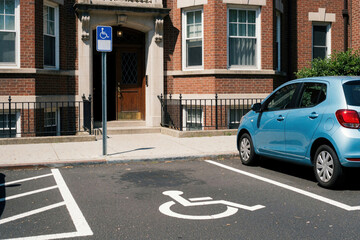 Fototapeta premium Blue car parked in accessible parking space near brick residential building entrance. Handicap symbol painted on asphalt with blue sign on pole. Disabled parking lot allows easy access for people