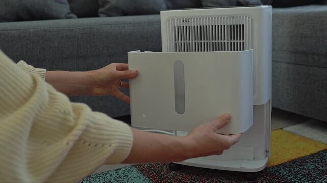 A woman removes the water tank from a dehumidifier, showing how much moisture was collected from a single room. Home appliance guides, moisture control articles, indoor air quality features, practical