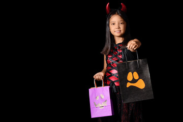 Fototapeta premium Halloween trick or treat, happy girl in devil costume holding candy bags for holiday celebration and autumn festivities on a black background