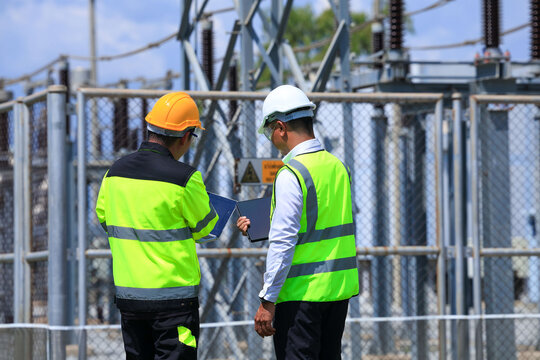 Electrical substation utility worker safety vest hard hat inspection engineer power grid industrial facility frame two technicians reviewing equipment behind secure fence on bright day