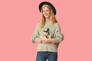 Fototapeta premium Autumn harvest and seasonal celebration, young woman holding decorative pumpkins for halloween festivities in a cozy sweater and hat on pink background
