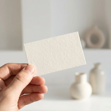 A hand holding a blank textured white business card in a minimalist interior setting.