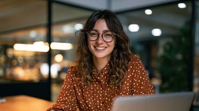 Portrait of a smiling young woman with glasses wearing a brown polka dot shirt while working on a laptop in a modern office environment