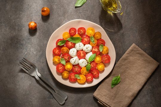 Italian salad of mozzarella, yellow and red cherry tomatoes and basil leaves in a plate on a dark grunge background, caprice salad, top view.