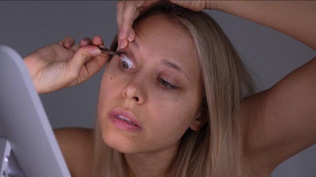 Young woman applies false lashes with tweezers in front of a mirror, demonstrating a detailed eyelash attachment process