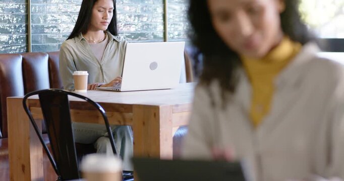 Asian woman working on laptop at coffee shop as camera shifting, right woman tapping tablet, cup