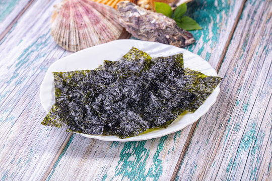 White ceramic plate holding dark green nori sheets beside scallop shell and oyster on weathered teal wooden surface, seaweed snack