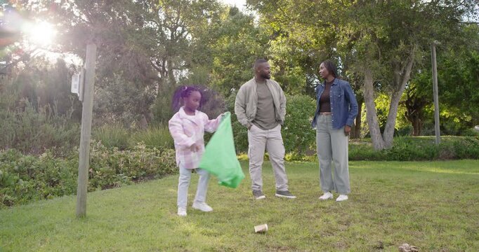 Family watching child spotting and picking up litter in park with green trash bag, parents smiling