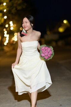 Elegant young woman in white dress holding pink flower bouquet at night outdoor