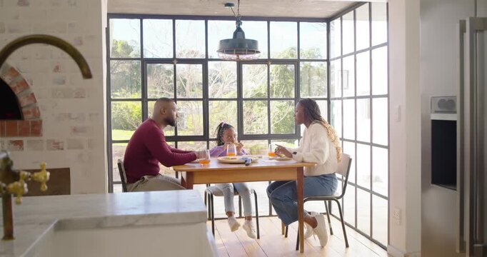 African American family feeding daughter during breakfast at table in nook, father passing glass