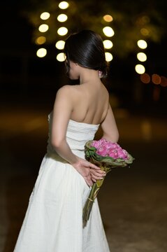 Woman in elegant white dress holding pink flower bouquet at night outdoor event