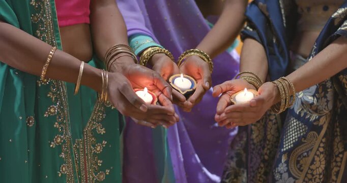 Three diverse women in saris reaching, holding central diya, lamps, steadying flames at home puja