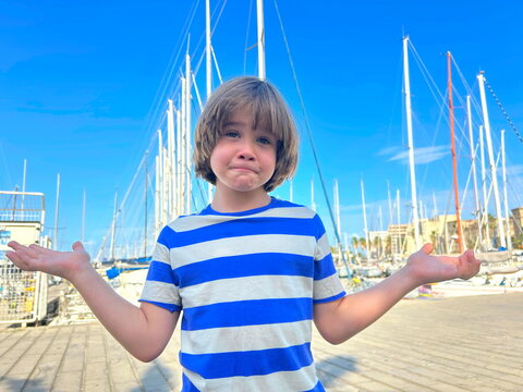 Portrait of a joyful smiling boy against a background of sailing boats. Yachting with children, family sea voyage. A child in a sailor t-shirt, ready to take the steering wheel
