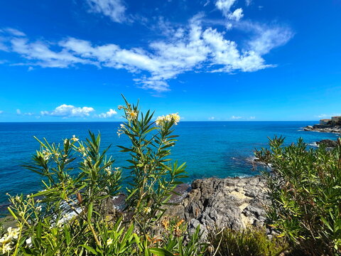 Scenic top-angle view of a Sicilian seascape with dramatic rocky cliffs and crags along the coastline. Crystal-clear turquoise Mediterranean waters crashing against the rugged shore under bright sunli