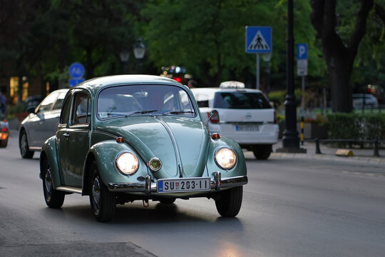 Vintage light green 1960s classic Volkswagen Beetle Type 1 early look car driving with headlights on along urban street, Subotica, Serbia, Jul 4, 2026