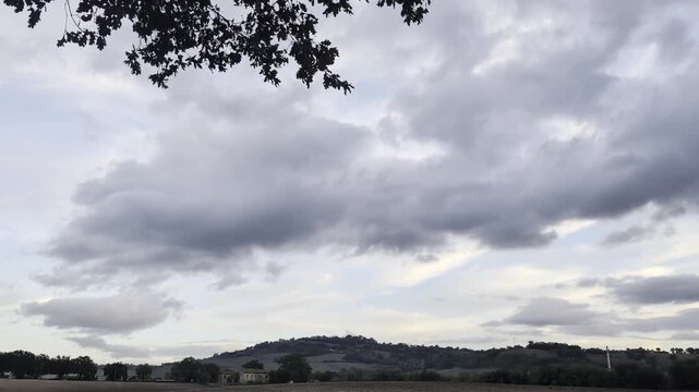 Linea collinare sotto un cielo nuvoloso e dinamico. Le grandi nuvole grigie e bianche si muovono lentamente sopra il paesaggio rurale, mentre le fronde di un albero incorniciano delicatamente la parte
