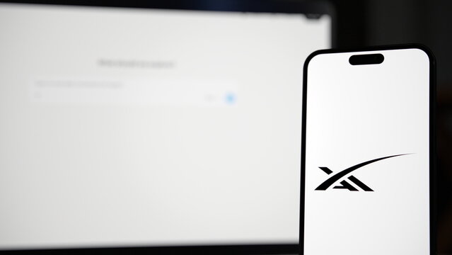 The xAI logo displayed on a smartphone held in front of a laptop showing the Grok website, at night with city lights in the background. October 8, 2026, Lisbon, Portugal.