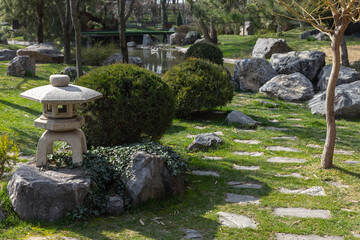 Fototapeta premium A serene Japanese garden scene featuring a traditional stone lantern beside stepping stones, pond reflections, rounded shrubs and natural rocks, evoking tranquility, meditation and landscape design
