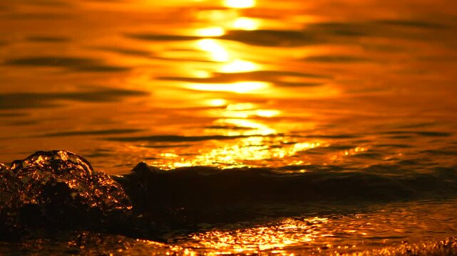 Golden sunset reflection glimmers on the gentle rhythmic ocean tides hitting a sandy shore. Steady eye-level close-up shot.