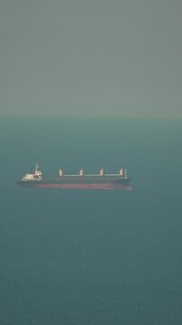 Bulk carrier transport ship glides across a tranquil blue ocean surface during a sunny day. Long distance telephoto shot.