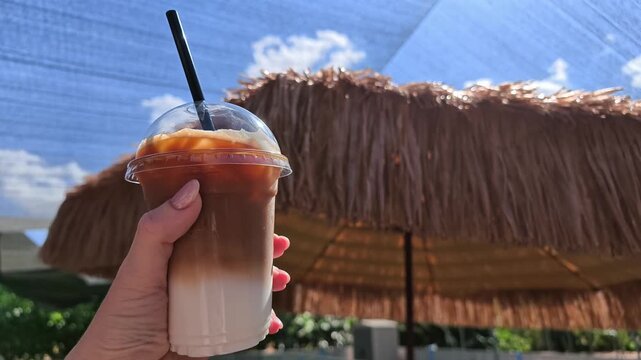 Hand holding an iced latte in a clear plastic cup with a domed lid and black straw, featuring distinct layers of milk and coffee against a thatched beach umbrella and blue sky