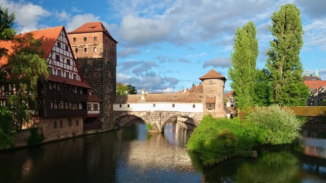 Real-time footage of the historic Henkersteg covered bridge, the Weinstadel and the Wasserturm reflected in the calm Pegnitz river under a blue sky in Nuremberg, Germany.