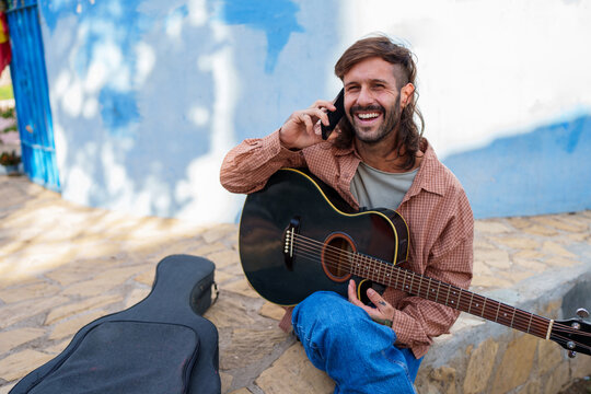 Male musician chatting on a cell phone, smiling and appreciating a peaceful moment with his acoustic guitar outside