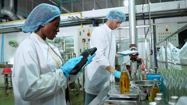 Two workers in protective clothing inspect bottles and monitor machinery during a hygienic beverage packaging process inside a modern industrial factory.