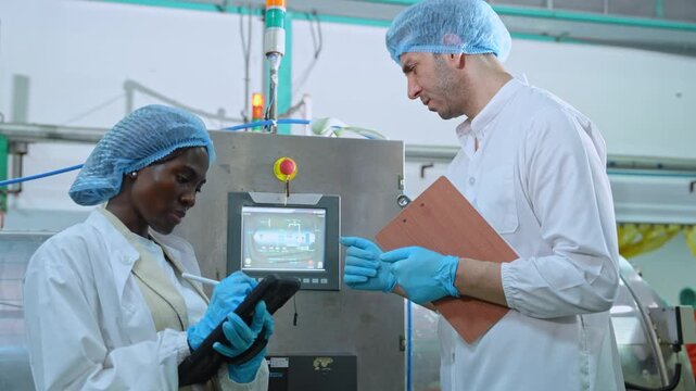 Two food production technicians in hygienic protective clothing inspect automated machinery and review quality-control data on a tablet inside a modern beverage packaging factory.