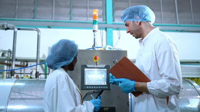 A production worker in protective clothing inspects an industrial control panel and records quality data inside a modern beverage processing and packaging facility.