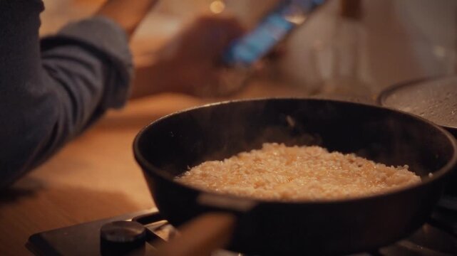 In home kitchen, cook attentively waits for rice to cook on stove. He glances at his smartphone, likely checking dinner recipes or responding to messages, as the aroma of cooking rice fills the air