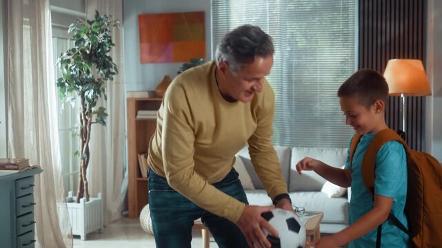 Grandfather helps his grandson put on a backpack, then they pick up a football and leave home together for practice, sharing a warm and supportive family moment.
