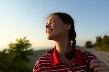 Fototapeta premium Smiling young woman with braided hair enjoys warm sunlight outdoors at sunset while wearing a striped polo shirt. Authentic portrait captures a happy, peaceful lifestyle moment in nature scenery.