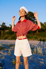 Naklejka premium A happy young woman in a striped shirt and shorts poses outdoors enjoying summer, lifestyle concept with casual clothes on a sandy beach during sunset, celebrating freedom and joy daily.