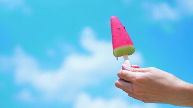 Close-up of hand holding melting watermelon popsicle dripping under bright blue summer sky. Hot weather, heatwave and refreshment lifestyle concept.