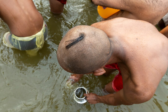 High angle view of a shaven headed novice with a topknot filling a steel cup with river water at Maha Kumbh Mela, Prayagraj, India