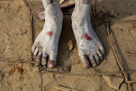 Close-up of bare feet of a Naga Sadhu covered in white ash with red marks on dry cracked ground at Maha Kumbh Mela, Prayagraj, India