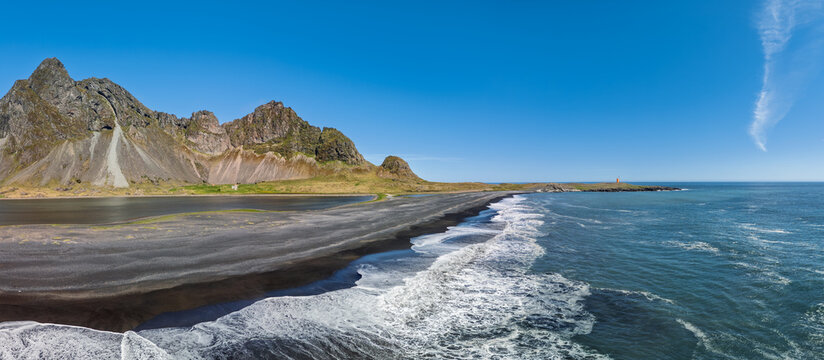 Panoramic aerial view of Hvalnes long black beach in Iceland, with Eystrahorn mountain, Lonsfjordur lagoon and white Atlantic surf beneath clear blue summer sky.