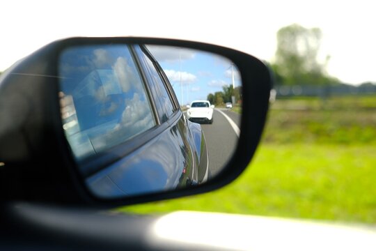 Close-up of a car side mirror reflecting a white car following behind on a country road under a blue sky with clouds
Side view mirror of a moving car