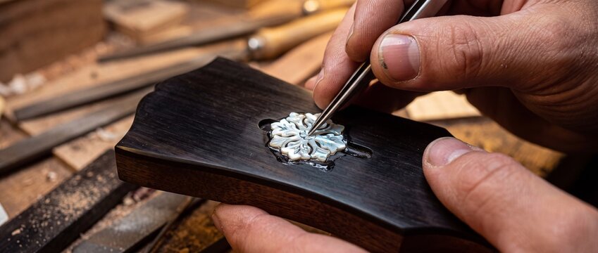 Close up view of tweezers placing intricate mother of pearl inlay onto dark ebony guitar headstock during high end instrument assembly