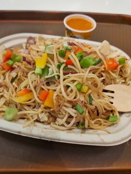 Veg hakka noodles with spicy red sauce, colorful bell peppers, capsicum, onions, cabbage, spring onions in eco friendly disposable paper plate with wooden fork, Indian Chinese street food top view.