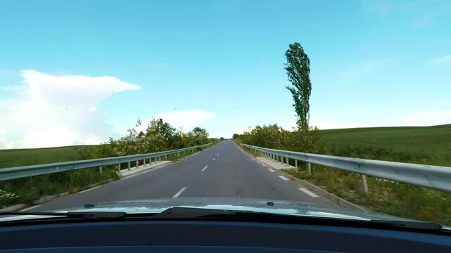 Windshield dashcam view with car hood driving along an open countryside road through green fields. Beautiful road trip on a sunny day under a blue sky with soft white clouds in Bulgaria.