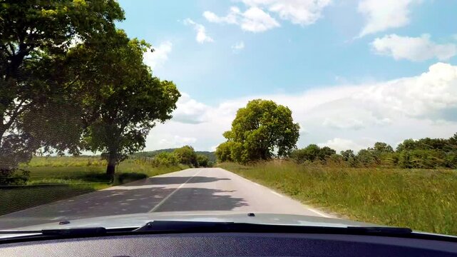Windshield dashcam view inside a car driving on a scenic countryside road lined with big green trees. Beautiful road trip on a sunny day under a blue sky with soft white clouds in Bulgaria.