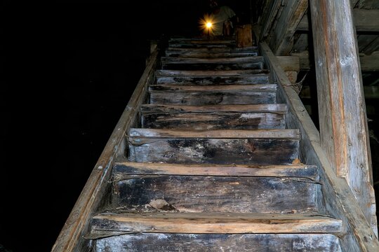 A person climbs a narrow stairway that fades into darkness. The stairs are worn and made of wood. A light source guides the way upward
