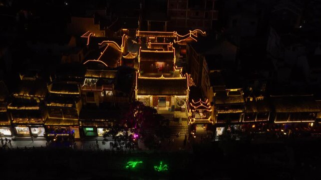 Slow pull-back from a historic hall with curved gable walls lit in gold, revealing the riverside promenade and neon characters at night, Fenghuang, Hunan, China