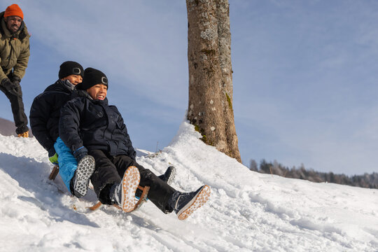 Happy family enjoying winter activities in nature, African American father pushing his two sons on a sled down a snowy slope.